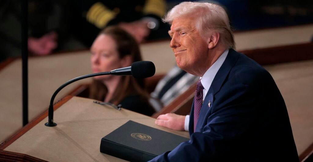 Donald Trump stands at a podium and looks to his left with a smirk on his face.