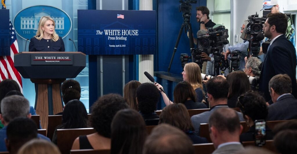 Karoline Leavitt stands at a podium while surrounded by members of the media who are seated in front of her.