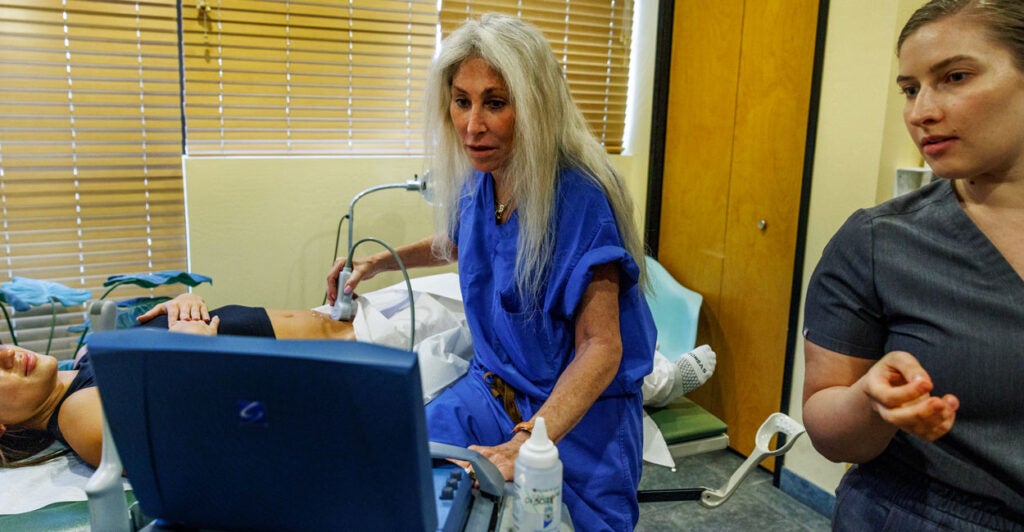 An abortionist in blue scrubs and long white hair performs an ultrasound on a patient with her nurse looking on.