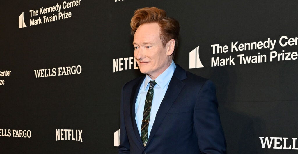 Conan O'Brien in a blue suit stands in a media photo op line