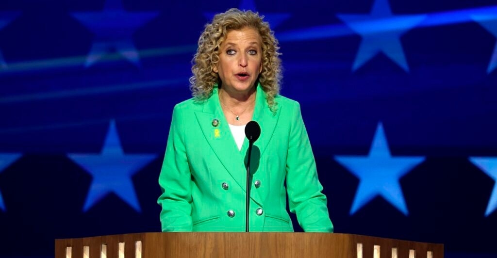 Debbie Wasserman Schultz stands at a podium in front of a microphone as she speaks at a conference.