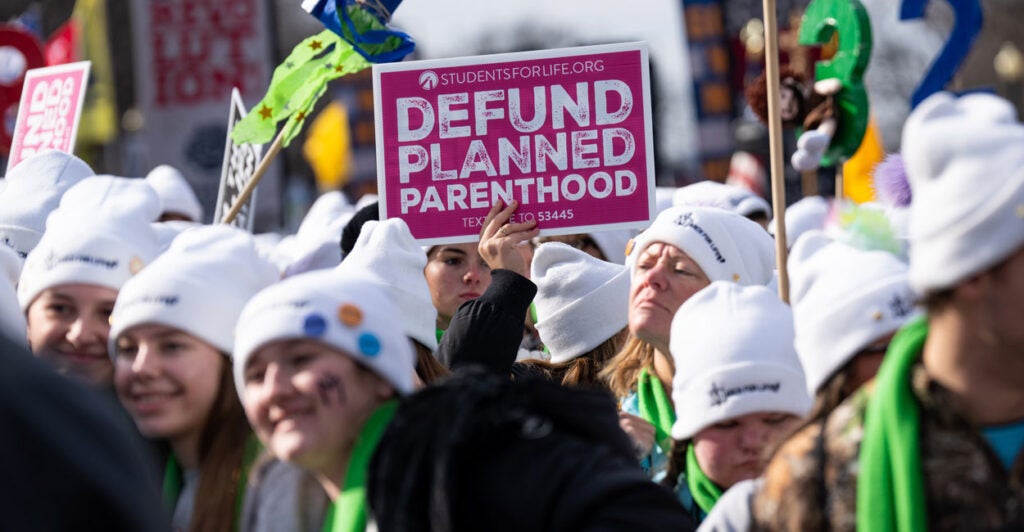 Protestors at the 2025 March for Life in white winter hats holding up Defund Planned Parenthood signs.
