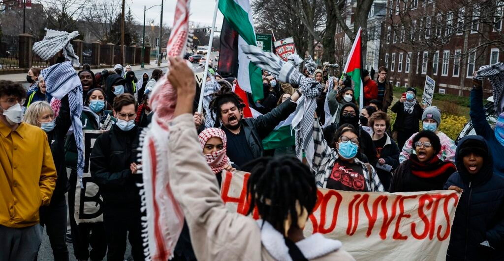 A group of students stand in the middle of the street and yell and wave flags as they protest.