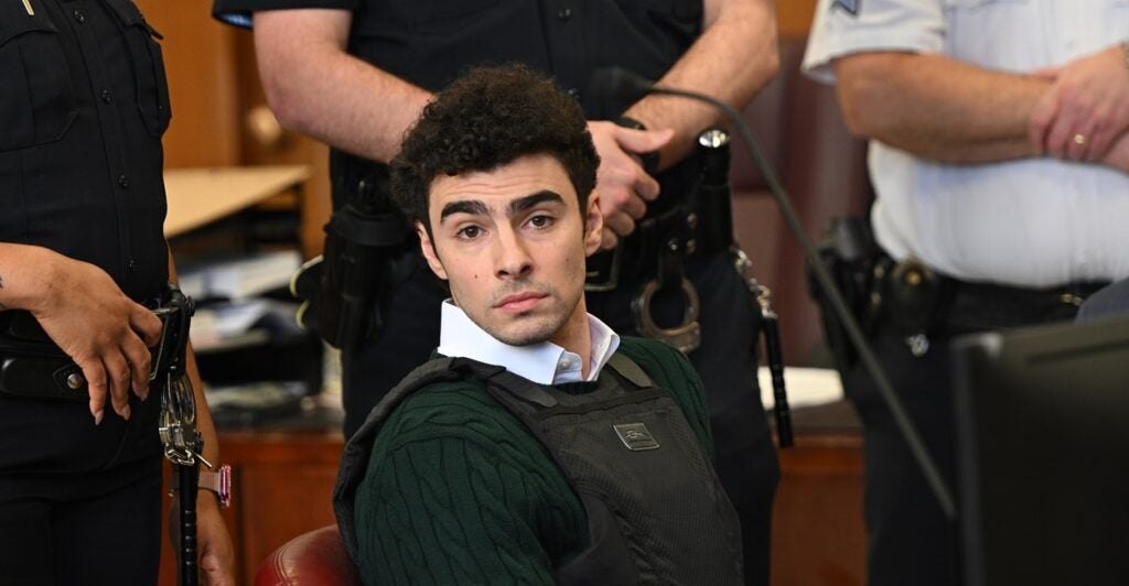 Luigi Mangione sits in a court room wearing a bulletproof vest surrounded by police as he looks to his right.
