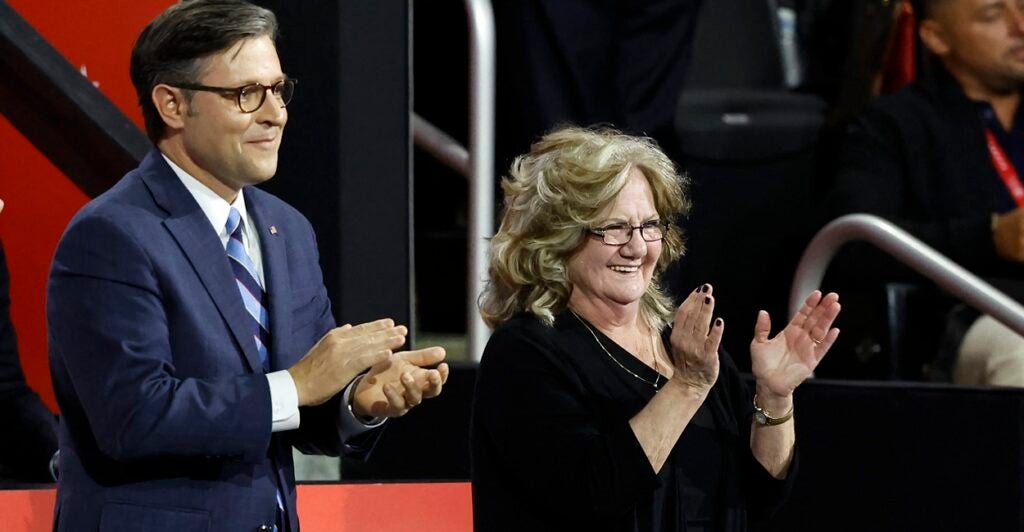 Bev Vance stands and applauds with a smile on her face as Mike Johnson stands to her right.