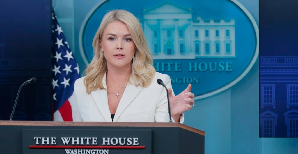 Karoline Leavitt stands behind a podium and holds up her left hand while talking to the press.