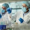 scientists in white robes and blue masks in a lab examining test tubes in beakers with liquids