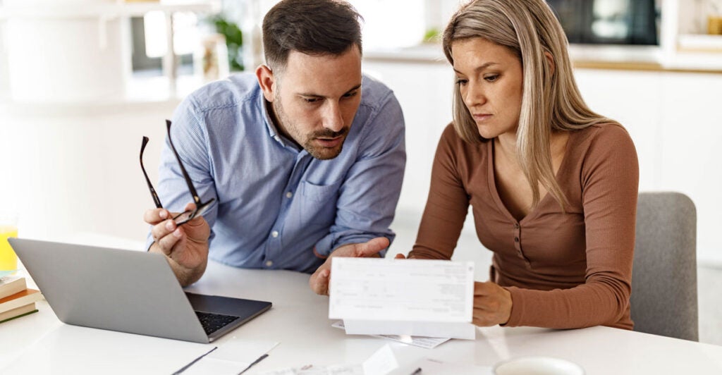 young couple sitting at the kitchen table with a laptop and tax paperwork looking worried
