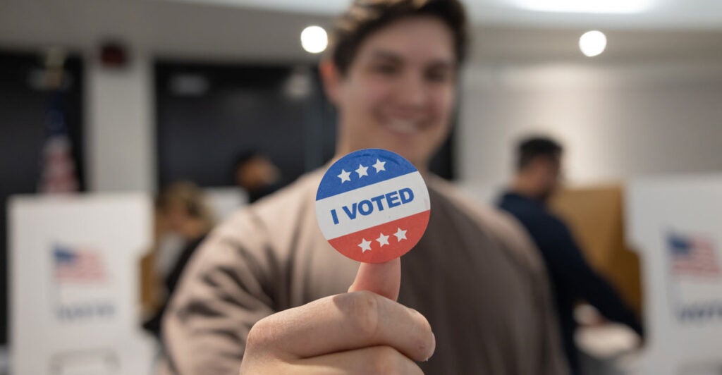 A 20-something man in the blurred background holding up an "I Voted" sticker.