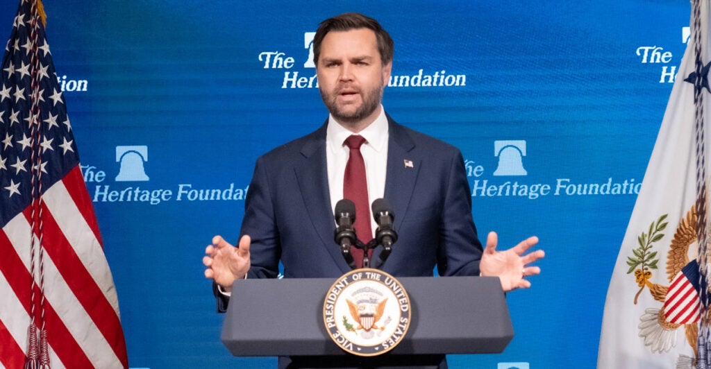 Vice President JD Vance in a blue suit and red tie at a lectern with microphones with an American flag in the background and the Heritage foundation logo in the background