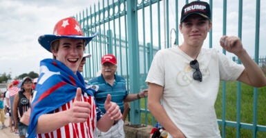 two young men wait to enter a Trump rally, one with a Trump hat on the other dressed in red white and blue with a Trump banner as a cape