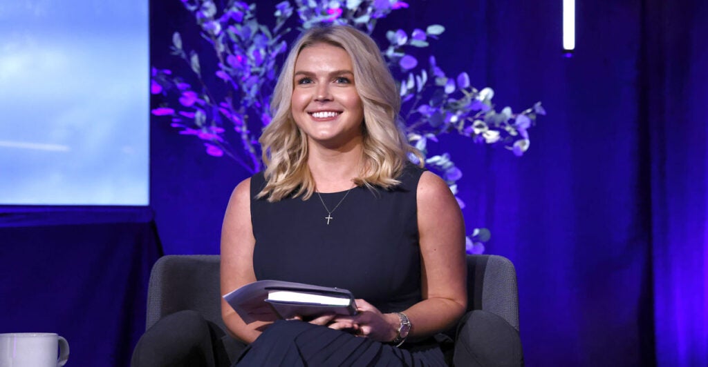 A smiling Karoline Leavitt in blue dress, in front of a backdrop of flowering tree.