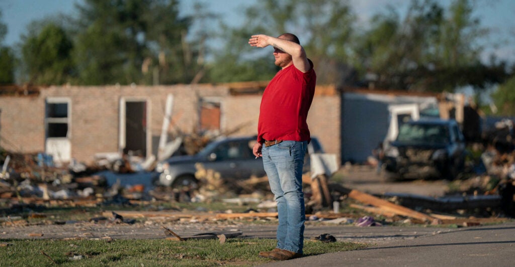 Man in red shirt and jeans with hand shielding his eyes, looks off, surrounded by tornado damage.