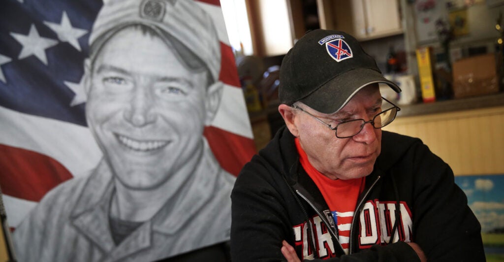 Gold Star father Paul Monti, arms folded, in front of a drawing of his smiling son Jared Monti.
