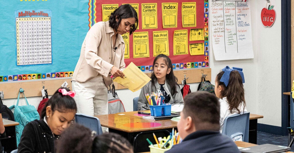 An Hispanic teacher stands over a student seated at a desk, showing her papers.
