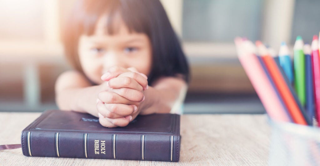 Brown-haired little girl with her hands clasped on top of a Bible. A bunch of crayons to her left.
