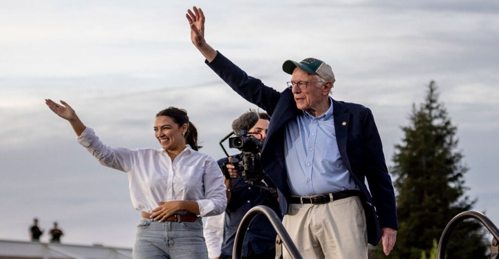 Bernie Sanders in blue jacket and hat, along with Alexandria Ocasio-Cortez in white shirt and jeans wave to crowd gathered for a "Fighting Oligarchy" rally.
