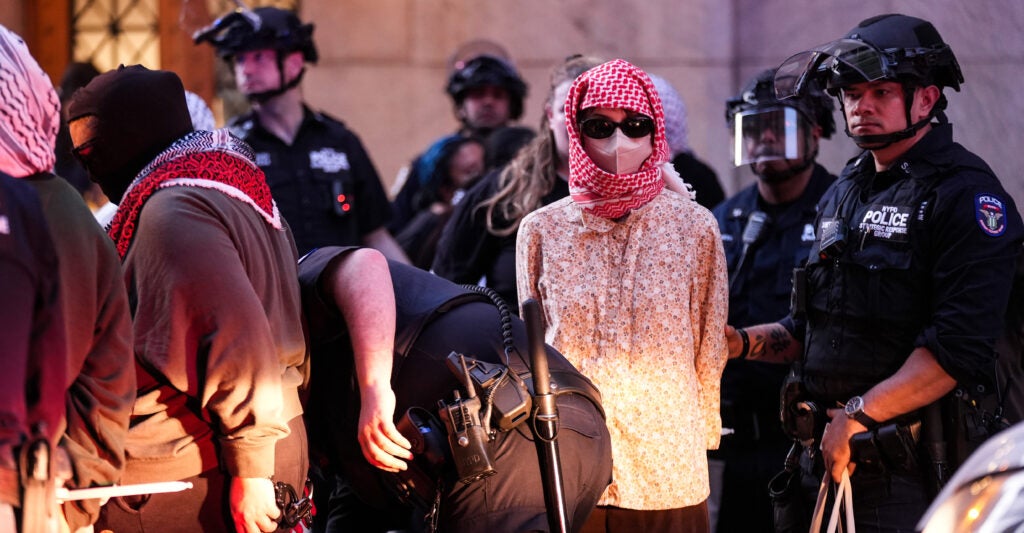 A masked protester in colorful blouse in police custody. Behind the suspect a row of heavily armored police. In front, an officer bends down.