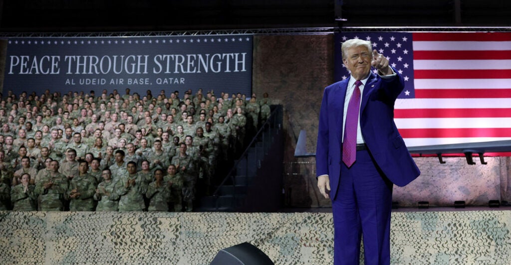 President Trump stands on stage at Al Udeid Air Base in a blue suit as troops look on.