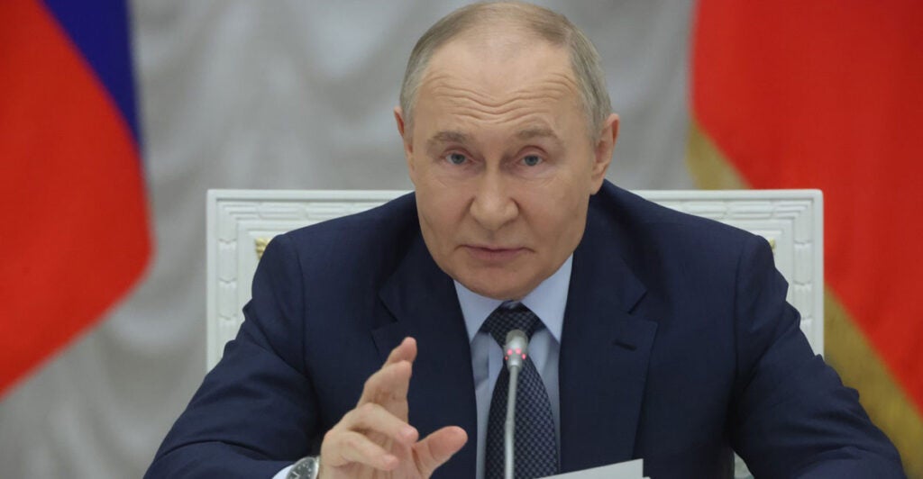 Vladimir Putin sits at a table behind a microphone and gestures with his right hand as he talks.