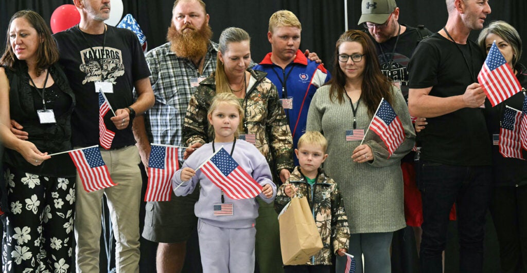a group of white Afrikaners, including men, women, and children, wave small American flags as they arrive in the United States for asylum