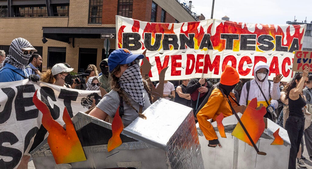 Protesters hold cybertrucks on fire and a sign reading "Burn a Tesla, Save Democracy"