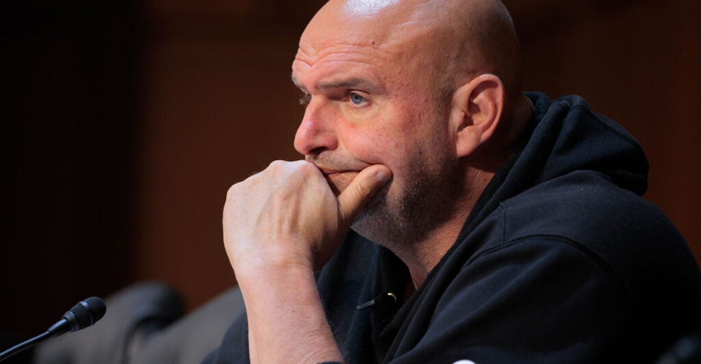 Senator John Fetterman sitting at a hearing in a hoodie with his chin resting on his hand