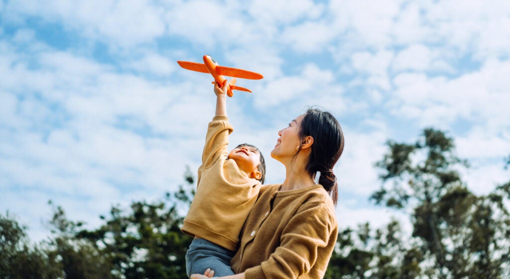 Young Asian mother and lovely little daughter spending time together outdoors, playing with airplane toy and smiling joyfully in park on a lovely sunny day against beautiful blue sky.