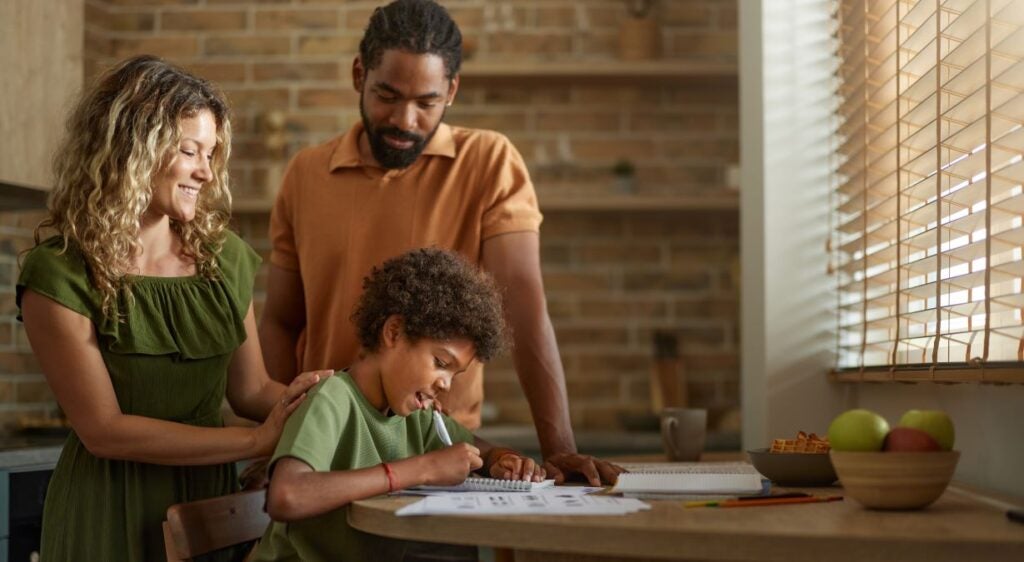 A black father and white, blonde mother affectionately stand over their son as he does homework.