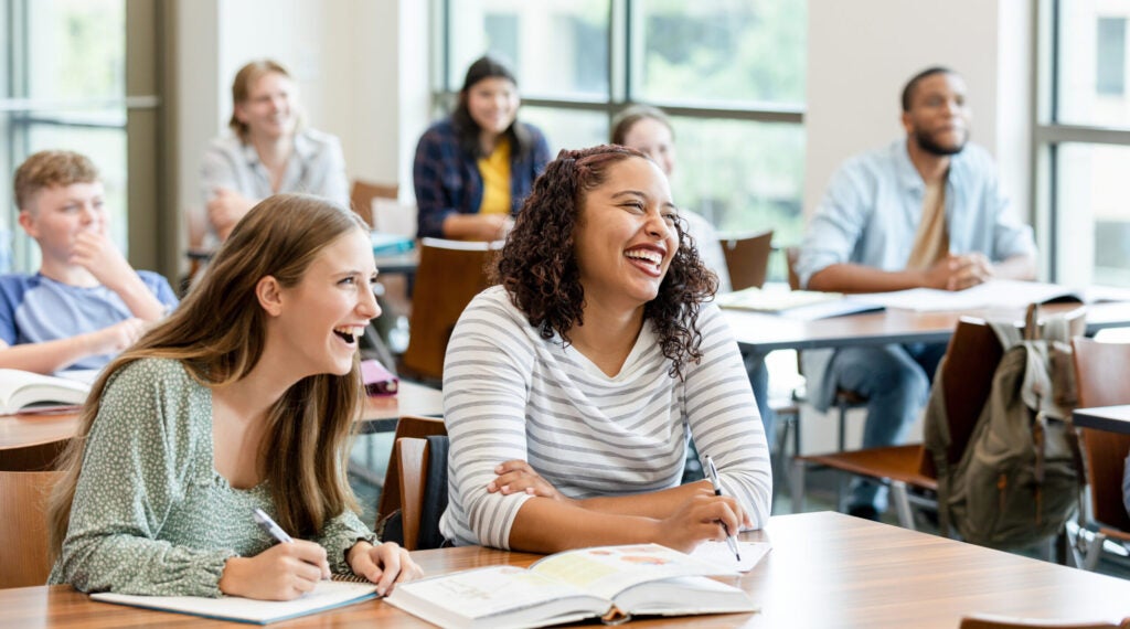 Two female college classmates at their desk laugh during a class. Four other students sit in background.
