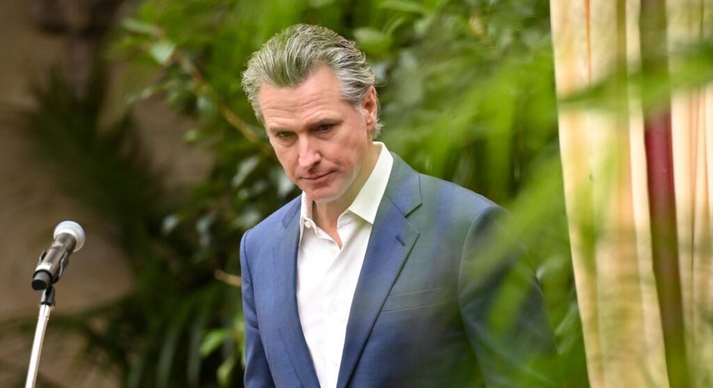 California Gov. Gavin Newsom, in a blue jacket and white open collar shirt, looks down and haggard, against a backdrop of green leaves.