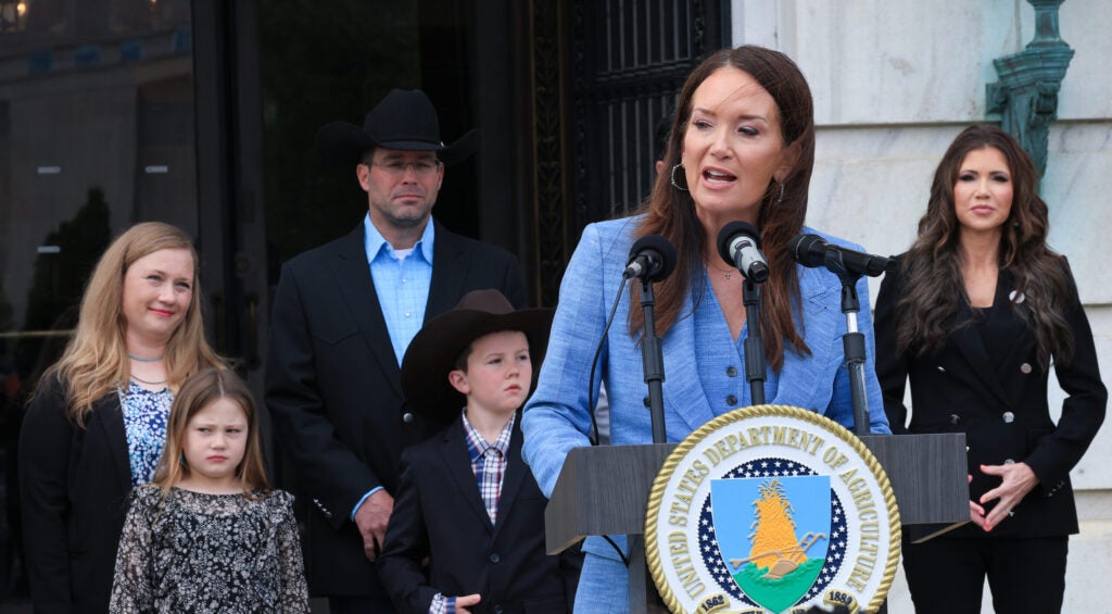 Agriculture Secretary Brooke Rollins in a blue outfit speaks at a podium, with DHS Secretary Kristi Noem and the Maude family behind her.