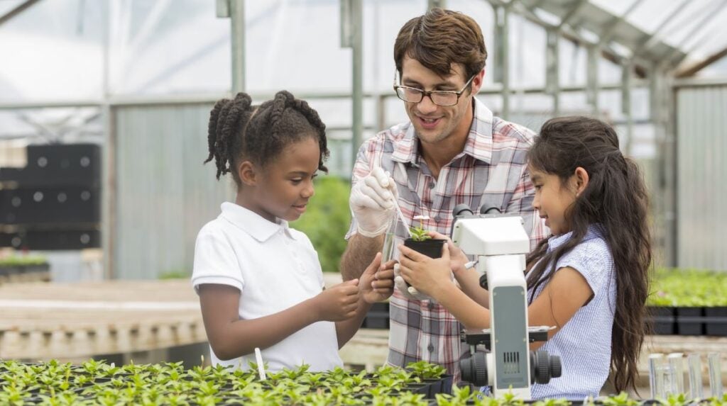 Inside a greenhouse, a white male botanist in glasses enthusiastically instructs a young black girl and young Hispanic girl. The Hispanic girl holds a small plant in a container that the botanist is probing.