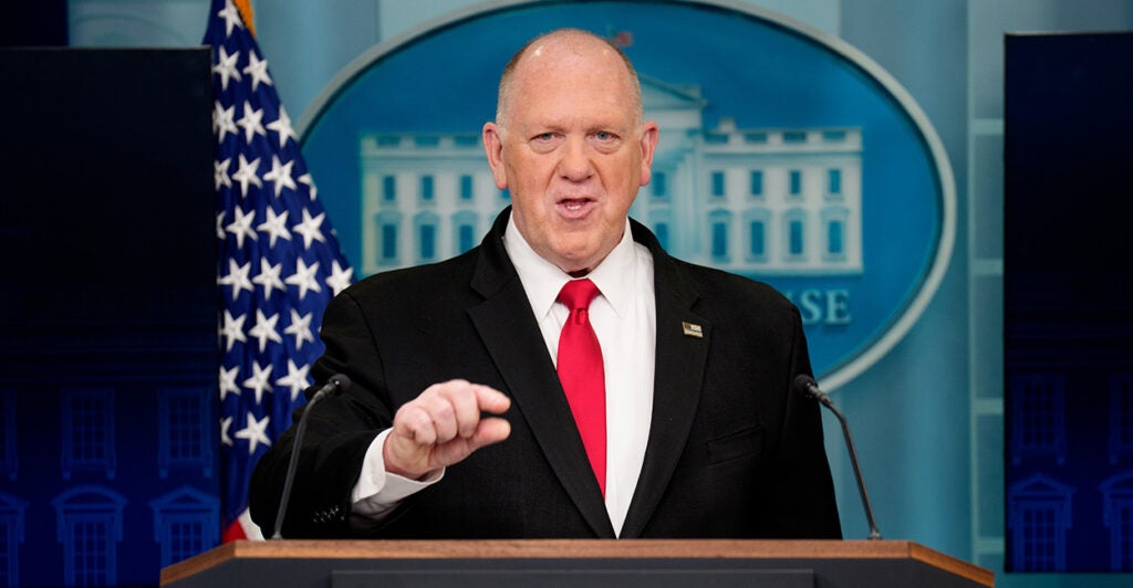 Tom Homan speaks from the podium of the White House press briefing room in a black suit and red tie.
