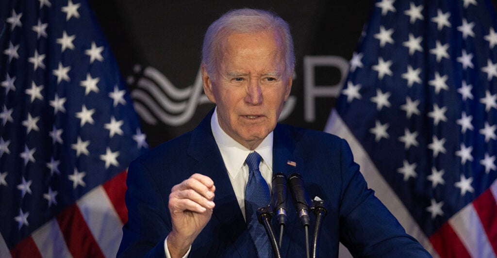 Joe Biden in a blue suit gestures in front of American flags