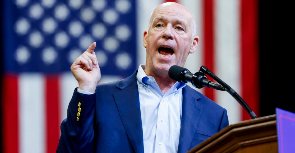 Montana Gov. Greg Gianforte speaking emphatically at a podium with hand raised in front of a large American flag backdrop