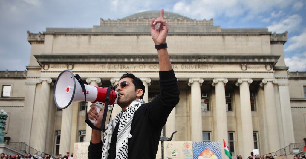 Mohsen Mahdawi in Palestinian garb with a megaphone and his hand raised in the air at a protest outside a Gothic structure stone building pillars on a university campus
