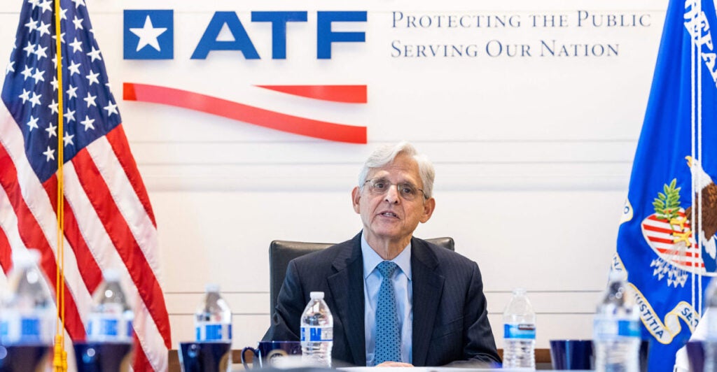 Then-U.S. Attorney General Merrick Garland in a dark suit and blue tie sitting at a conference table at the ATF with an ATF logo on the wall in back of him