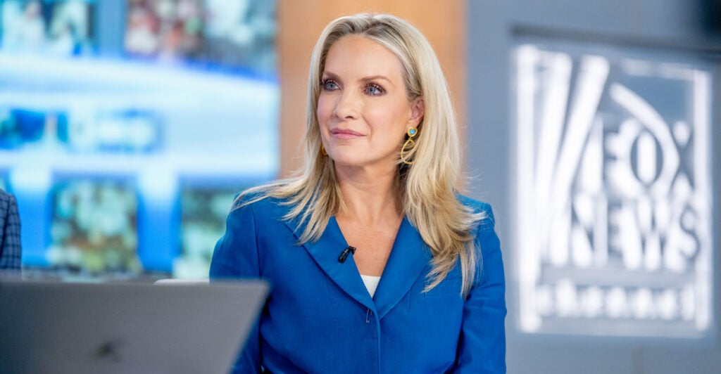 Fox News Host Dana Perino in a blue suit sits in studio at the Fox News desk with the Fox News logo on the wall behind her