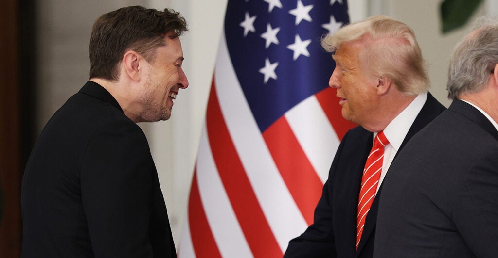 Elon Musk and President Donald Trump shaking hands in front of an American flag.