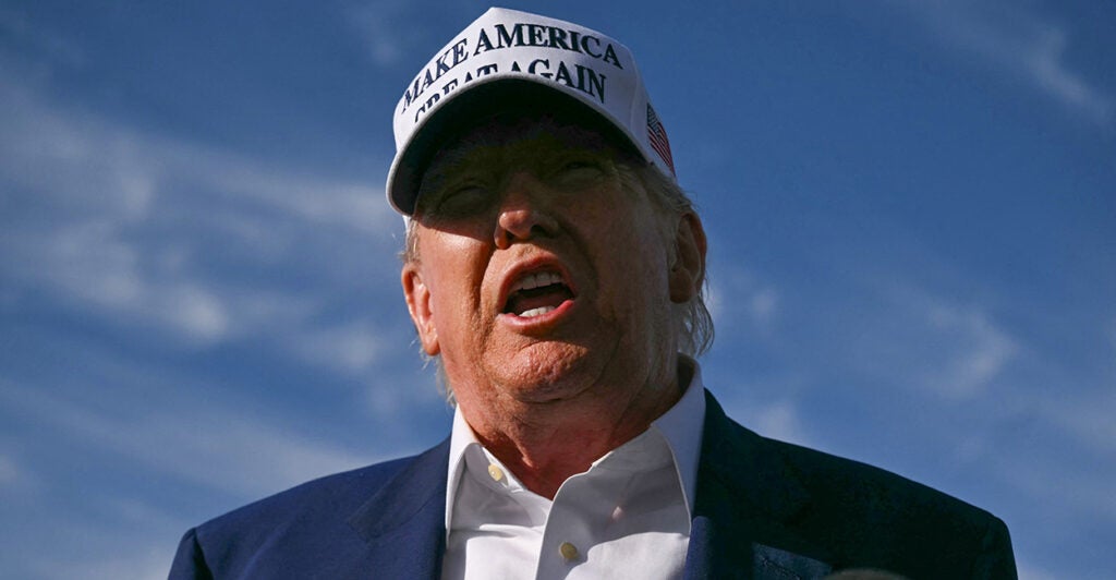 Close-up from a low angle of President Trump speaking while wearing a white "Make America Great Again" hat with blue lettering.