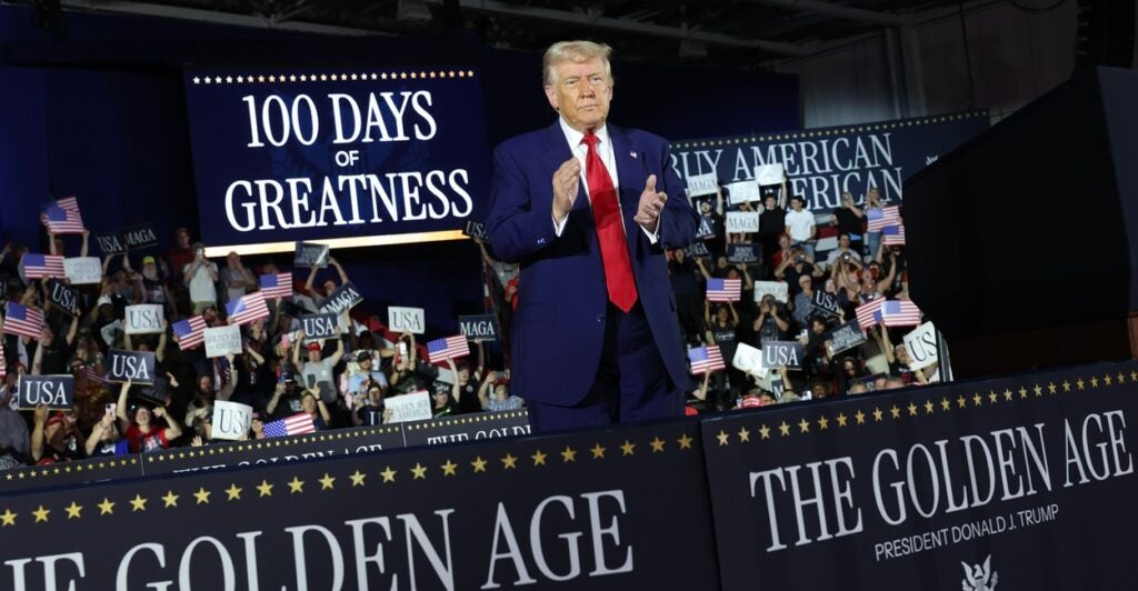 Donald Trump stands behind a banner and applauds while looking out at the crowd.