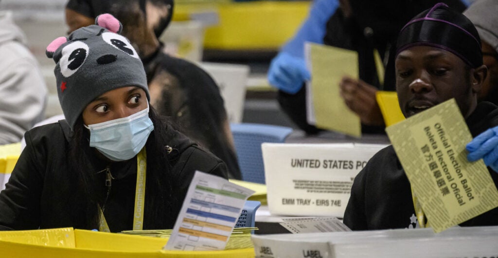 Two black ballot counters in Philadelphia. On the left, a wide-eyed woman with a mask and a mouse wool cap and on the right a male holding up a yellow ballot.