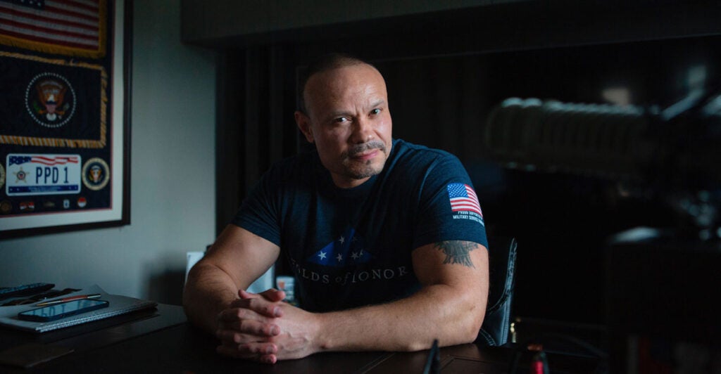 Dan Bongino, sits at a desk in his studio, in a dark blue"Folds of Honor" t-shirt.