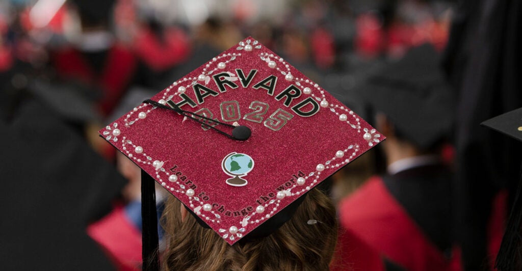 A crimson Harvard graduation cap atop the head of a graduate. The hat reads "Harvard 2025" with a tassel crossing to the left, and in small letters "Learn to Change the World."