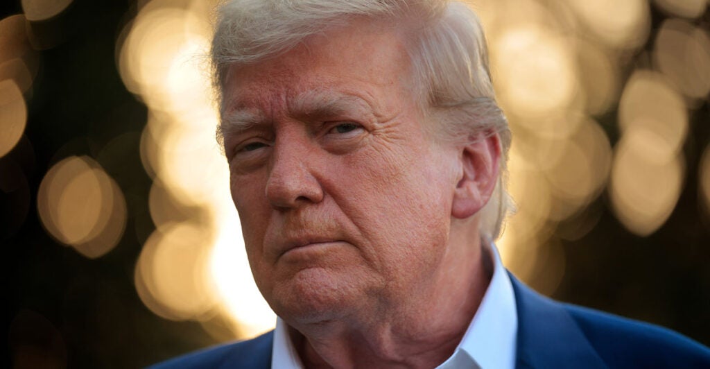 Close-up of Donald Trump in a blue jacket with a white open collar against a backdrop of diffused lights.