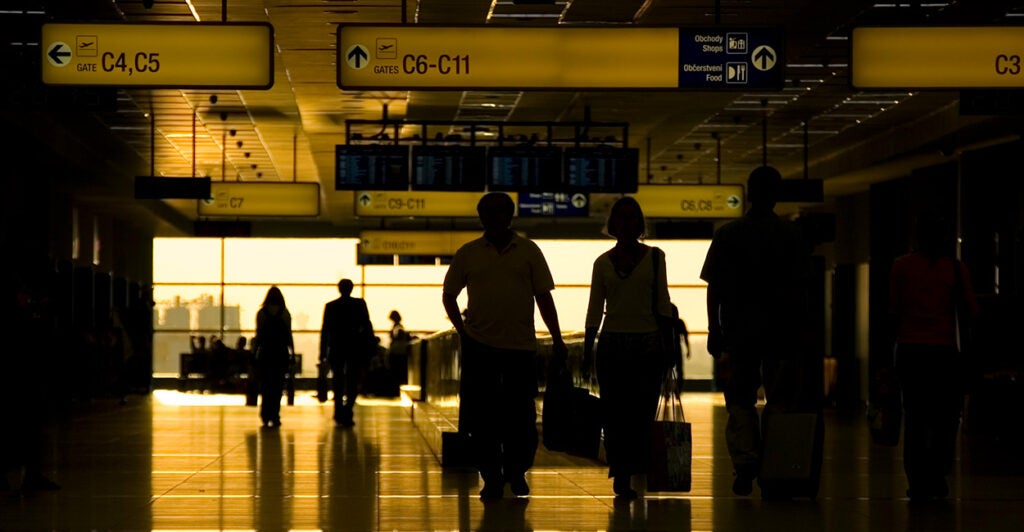 People are seen walking through an airport terminal in dark lighting.