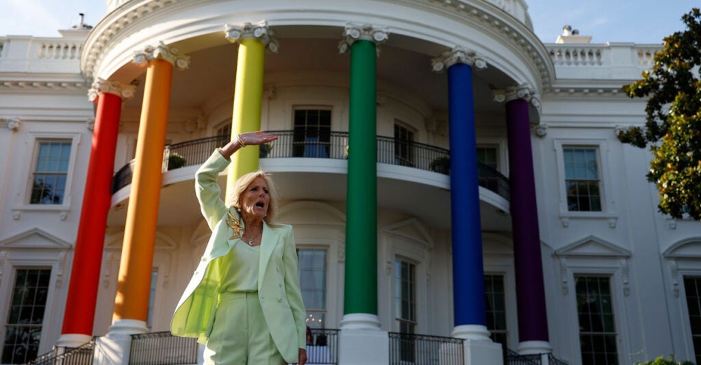 Jill Biden stands in front of the White House that has been decorated in rainbow colors while holding her right arm up in the air.
