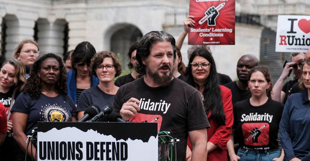 Todd Wolfson stands outdoors at a podium with a microphone and he turns his head to the left to look at the crowd.