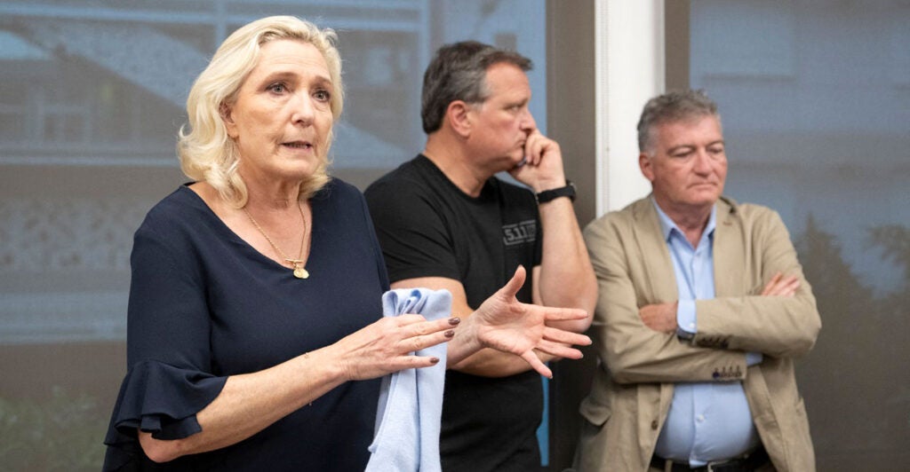 Marine Le Pen stands gesturing with her hands while talking to a crowd of reporters.